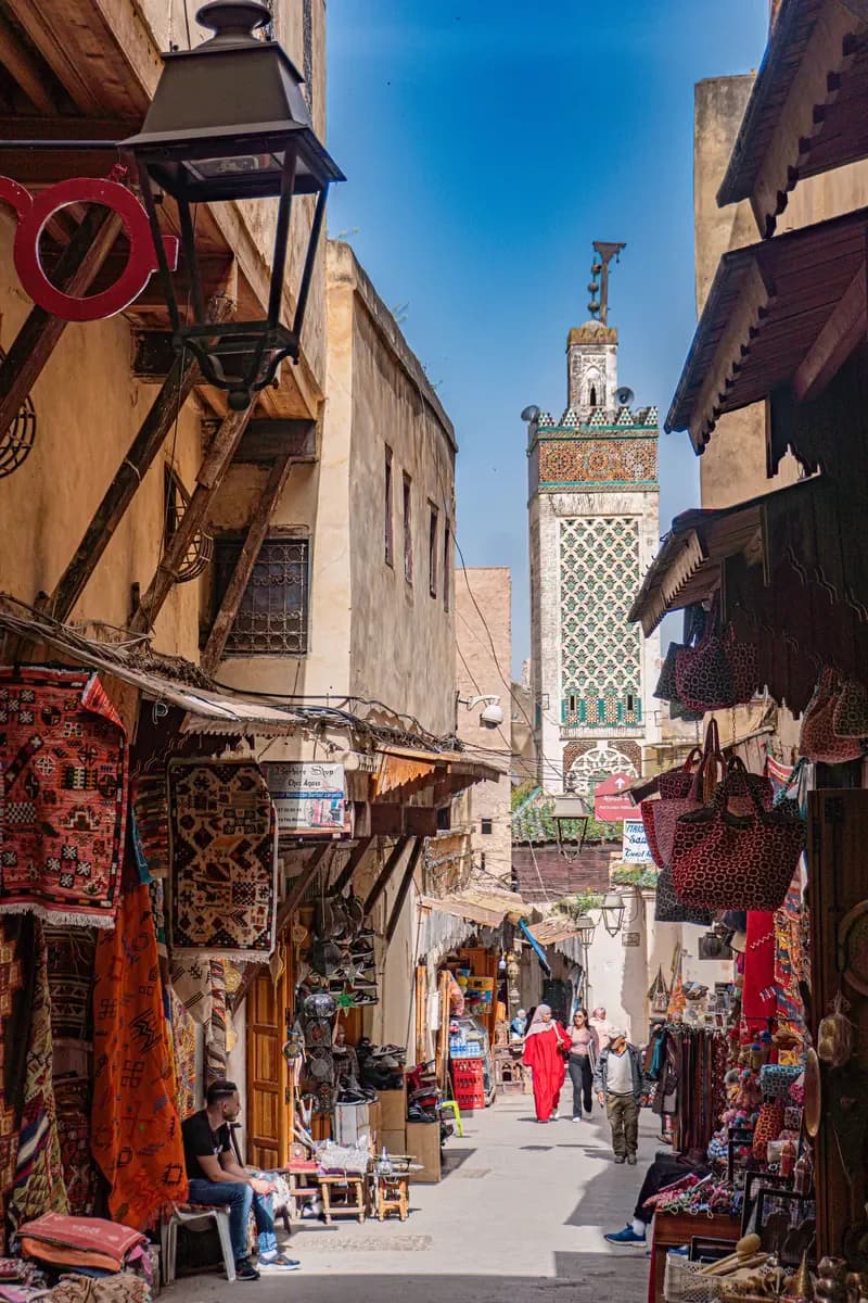 A souk lane in the Fes medina, rugs and bags hung outside the shops, a minaret ahead
